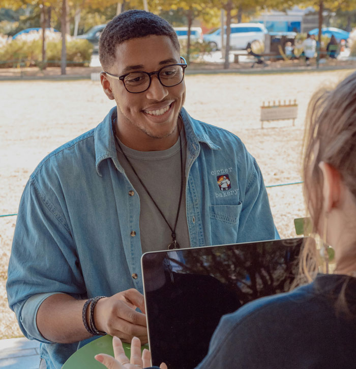 Smiling man in glasses and denim shirt, seated outdoors, engaging in conversation, embodying real-life quality hacks.