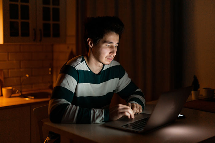 Man in striped shirt using laptop alone at night, symbolic of friend group deception.