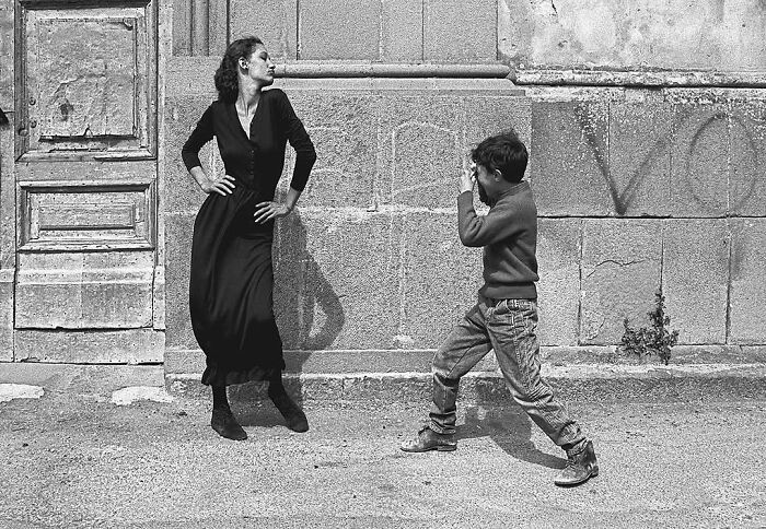 A woman posing confidently as a young boy playfully pretends to photograph her against a stone wall; interesting images theme.