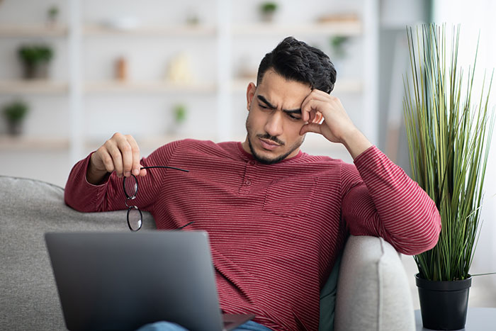 Man frustrated over apartment scam, holding glasses, looking at laptop in living room.