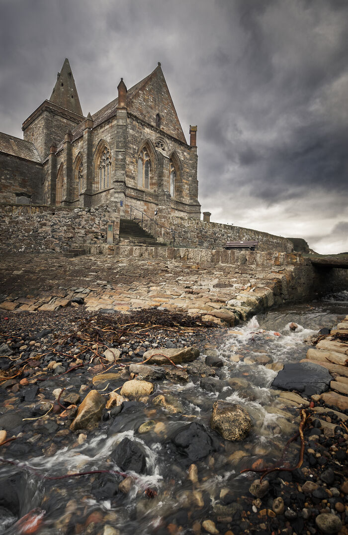 Gothic-style church by rocky stream under dramatic sky, showcasing fascinating church architecture.