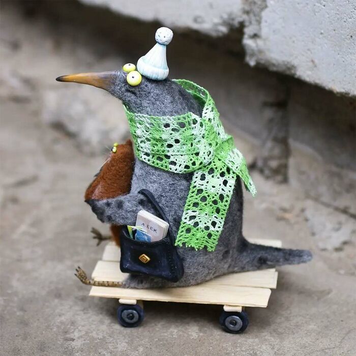 Funny-looking animal doll by Georgian artist, wearing green lace scarf, holding a bag, perched on a wheeled platform.