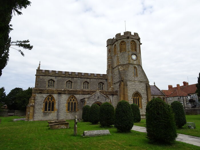 Ancient stone church with clock tower and arched windows, surrounded by neatly trimmed bushes under a cloudy sky.