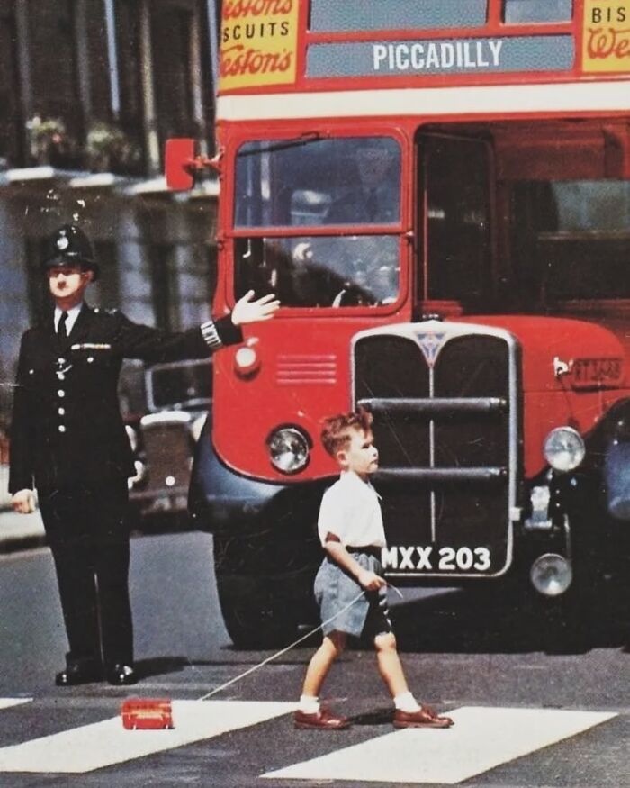 Child with toy, crossing street escorted by police officer, interesting image of vintage London scene with double-decker bus.