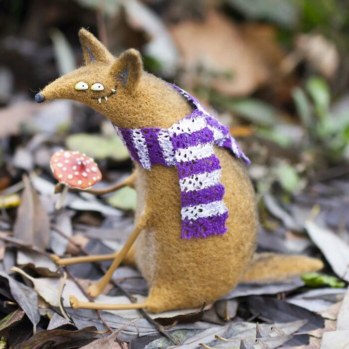 Funny-looking animal doll in a purple scarf, crafted by a Georgian artist, sits among leaves with a mushroom nearby.