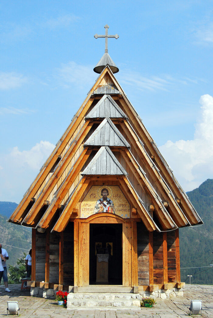 Wooden church with a cross on top, set against a clear sky, surrounded by mountains.