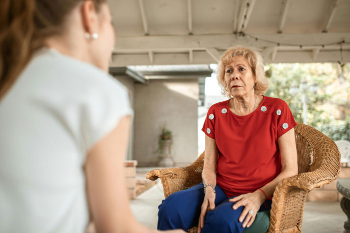 Woman in a red shirt sitting in a wicker chair, looking concerned, talking to someone off-camera.