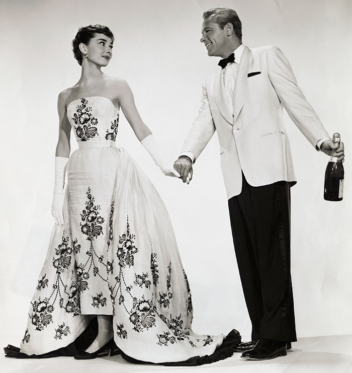 A woman in an elegant, embroidered gown and gloves holds hands with a man in a tuxedo, exuding elegance and class.