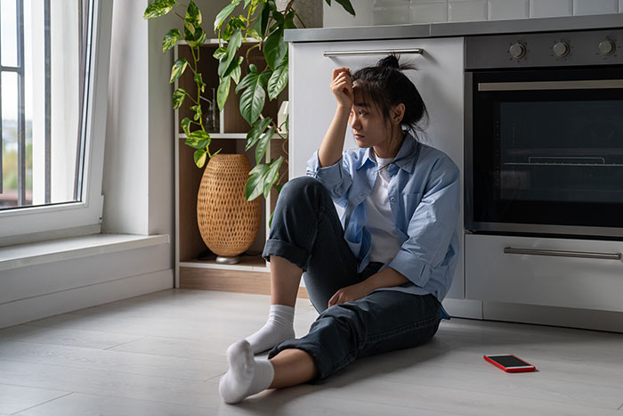 Woman looking shocked, sitting on kitchen floor beside a plant, reflecting on boyfriend\'s fictitious friend group.