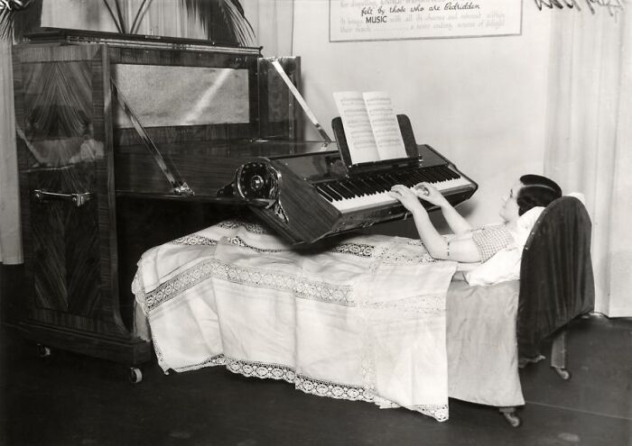 A woman plays a horizontal piano while lying in bed, an innovation ahead of its time.