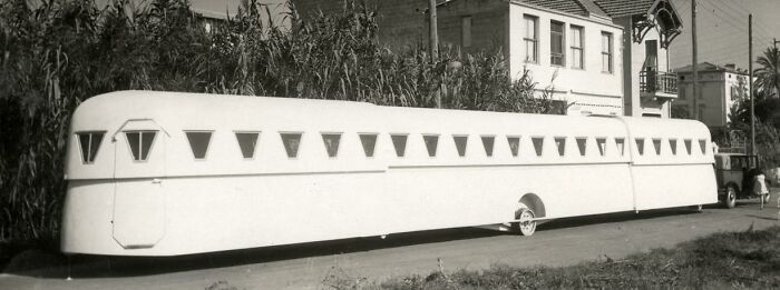 Long vintage streamlined bus, a historical invention ahead of its time, parked on a street with buildings in the background.