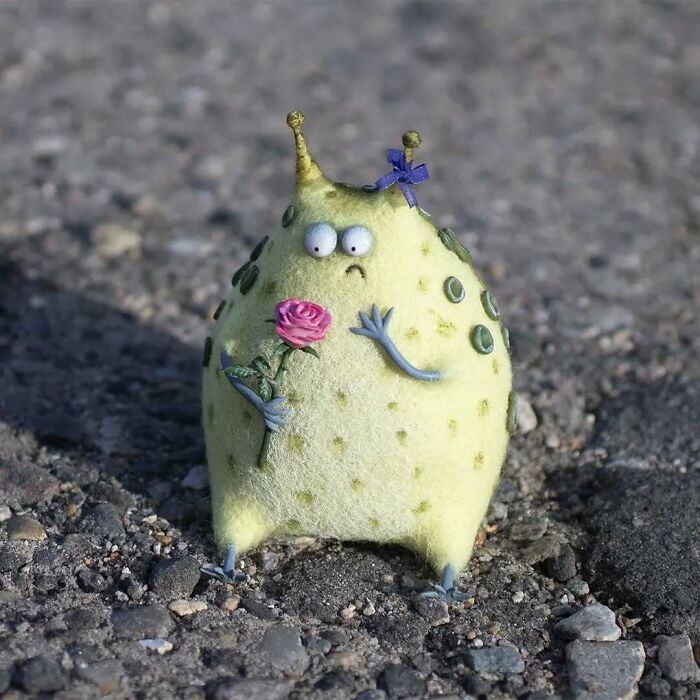 Funny-looking art doll resembling an animal, created by Georgian artist, holding a pink flower on a rocky surface.