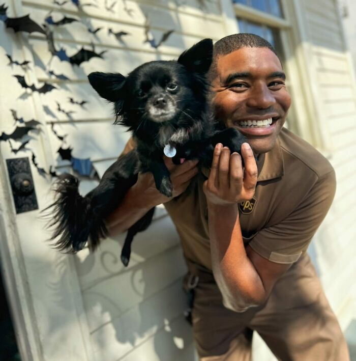 UPS driver smiling while holding an adorable black dog near a decorated wall.