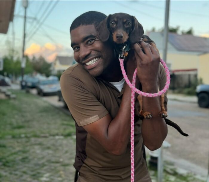 UPS driver smiling with a small dog on shoulder, pink leash in hand, bright street setting.