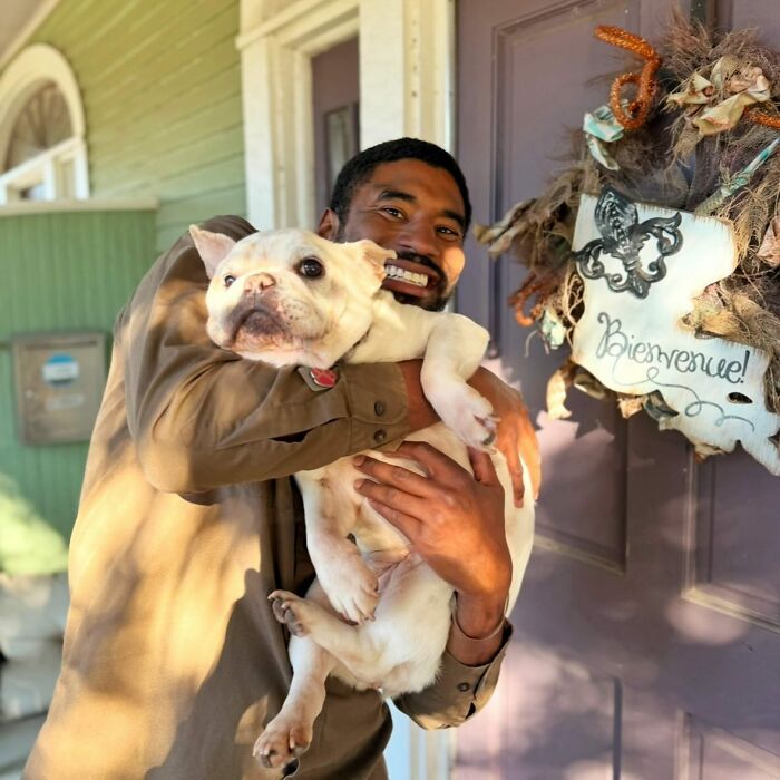 UPS driver holding a cute bulldog outside a home with a decorative wreath on the door.