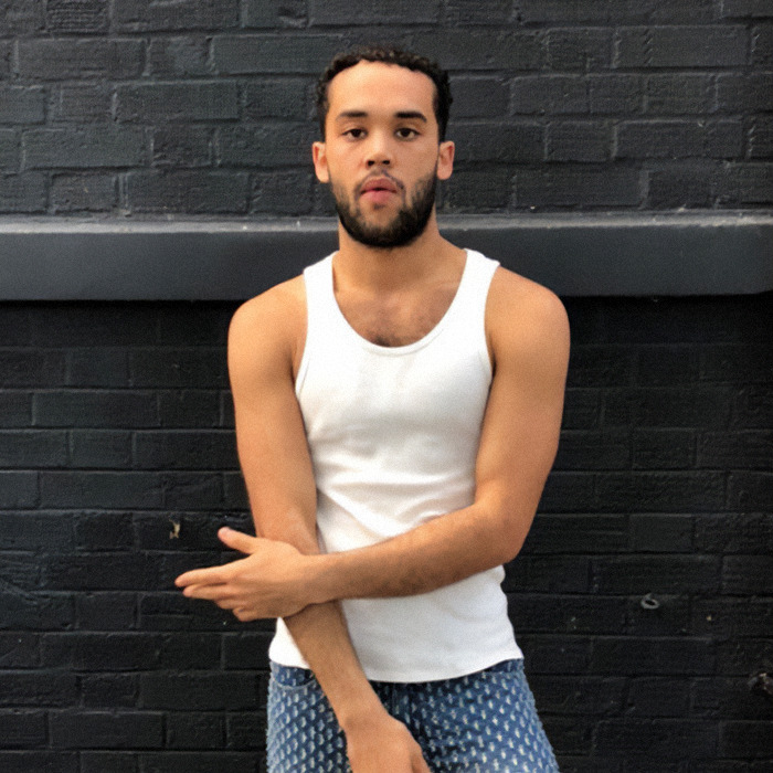 Kanah Flex in a white tank top against a black brick wall.