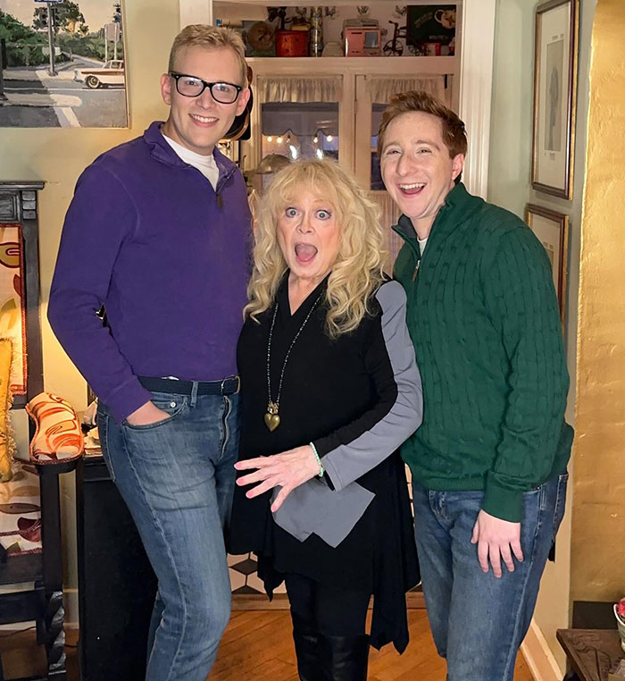 Three people, including a woman with long blonde hair in the center, smiling for a photo in a warmly lit room.