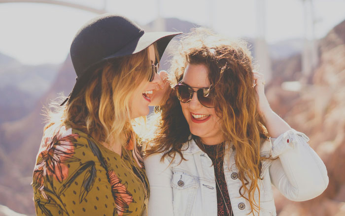 Two women smiling in sunlight, wearing sunglasses and casual outdoor clothing, enjoying a moment together.