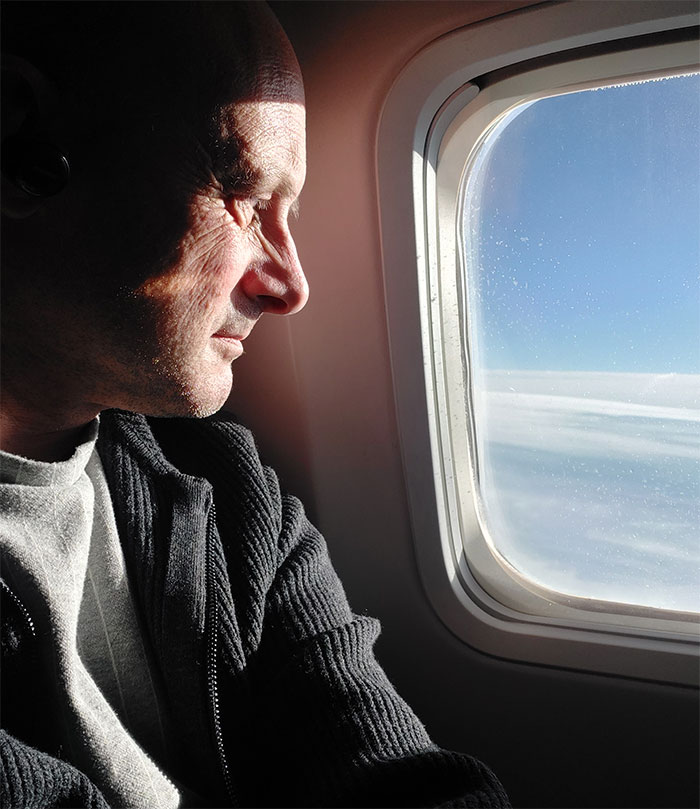 Man looking out airplane window, sunlight on face, reflecting on stubborn boomer's row-by-row deplaning insistence.