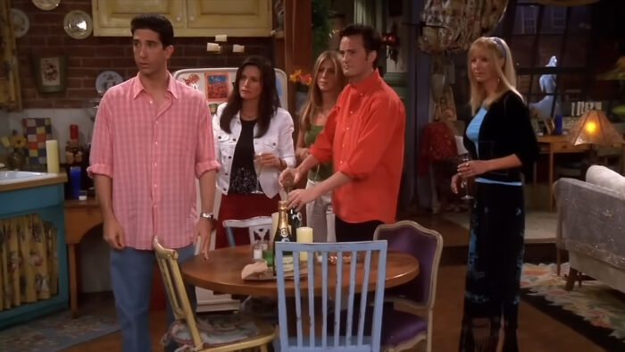 Five people standing around a kitchen table with wine glasses, from a beloved show known for comedy.