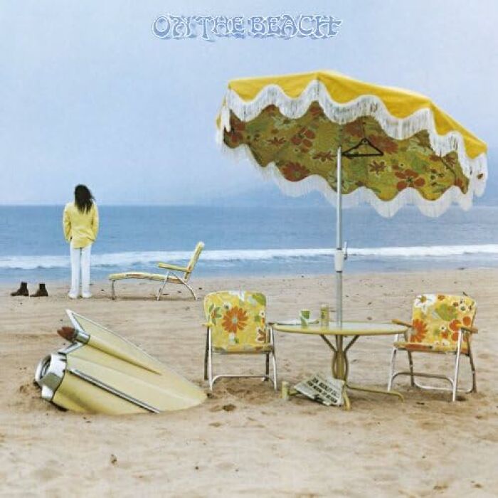 Beach scene with vintage '70s furniture, a standing person by the ocean, and a floral umbrella.