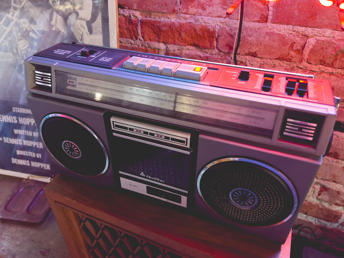 A vintage 80s boombox with cassette deck and speakers, placed on a wooden surface against a brick wall.