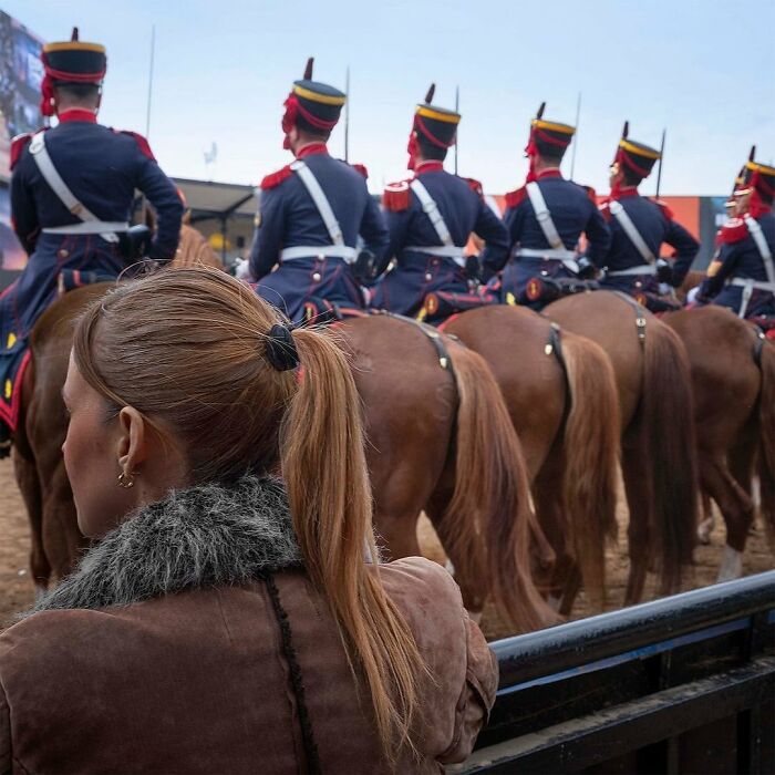 A woman watches soldiers in ceremonial uniforms on horseback in a parade setting.