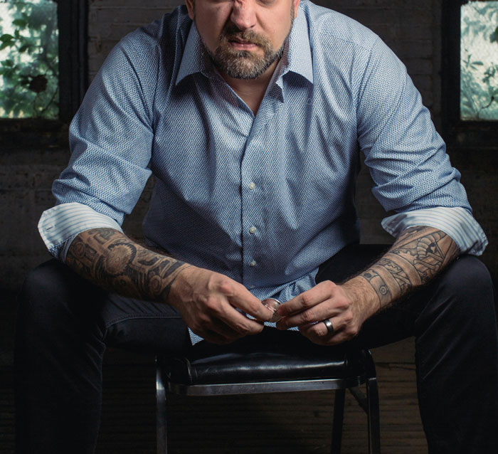 Man with tattoos sitting on a chair in a dimly lit room, conveying a "we don’t talk about that" family story atmosphere.