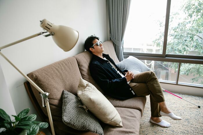 Man lounging on a sofa, wearing sunglasses, with a relaxed posture next to a window; represents a casual unethical life hack.