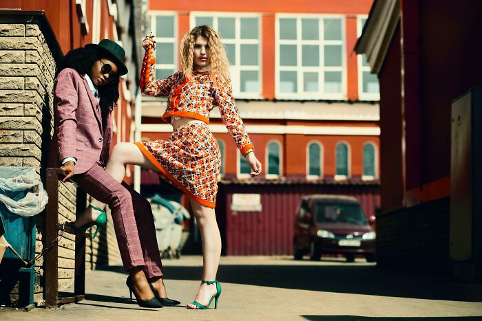 Two stylish women posing in an alley, one in a pink suit, the other in a patterned outfit, showcasing fashion trends.