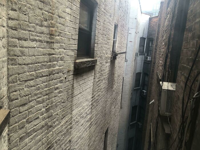 Narrow, grimy alley between tall brick buildings representing urban hell conditions with AC units and small windows.