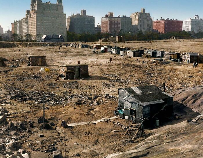 Urban hellscape depicting a barren area with makeshift structures, contrasting with distant city buildings.