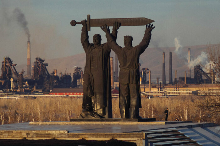 Statues holding a sword in an urban hell scene with industrial smokestacks in the background.