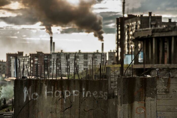 Urban hell scene with smoke from factories, crumbling buildings, and graffiti reading "No Happiness" on a concrete wall.