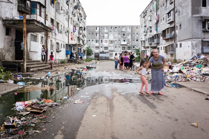 Urban hell scene with rundown apartments, litter-strewn streets, puddles, and people walking through the area.