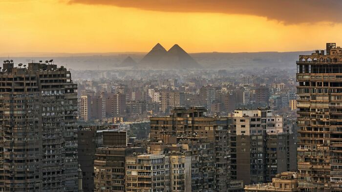 Urban hell: Dense cityscape with old buildings, pyramids in the background, under a dramatic orange sky.