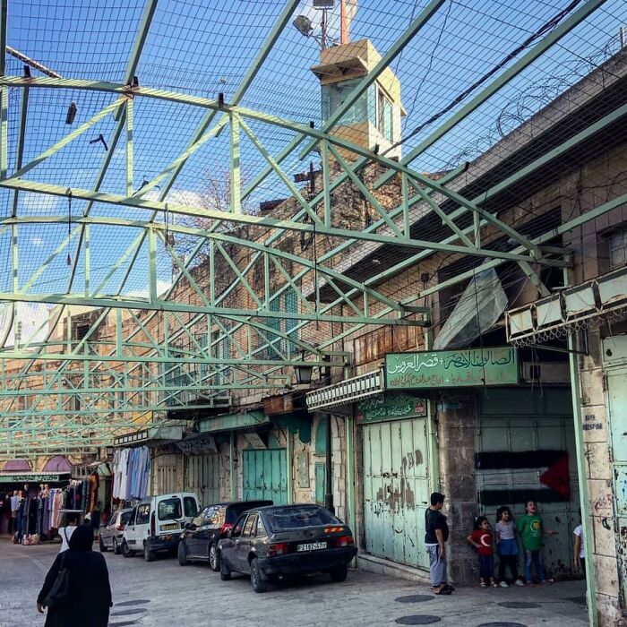 Urban decay scene with metal netting above, worn buildings, parked cars, and people walking, reflecting urban hell.