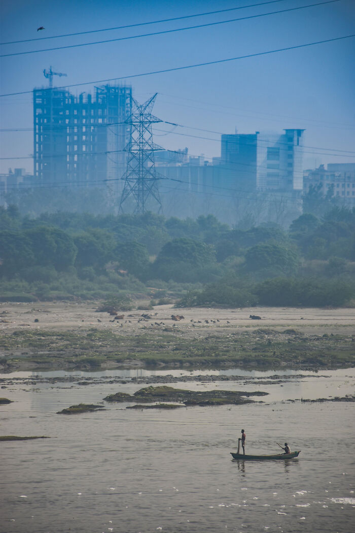 Urban hell example: polluted river with two people in a boat, power lines, and a hazy cityscape in the background.