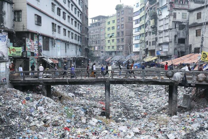 People walking on a bridge over a trash-filled area between rundown buildings, depicting an example of urban hell.