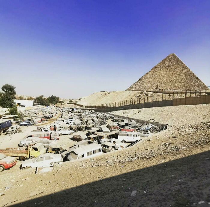 Urban hell scene with a car junkyard near a large pyramid under a clear blue sky.