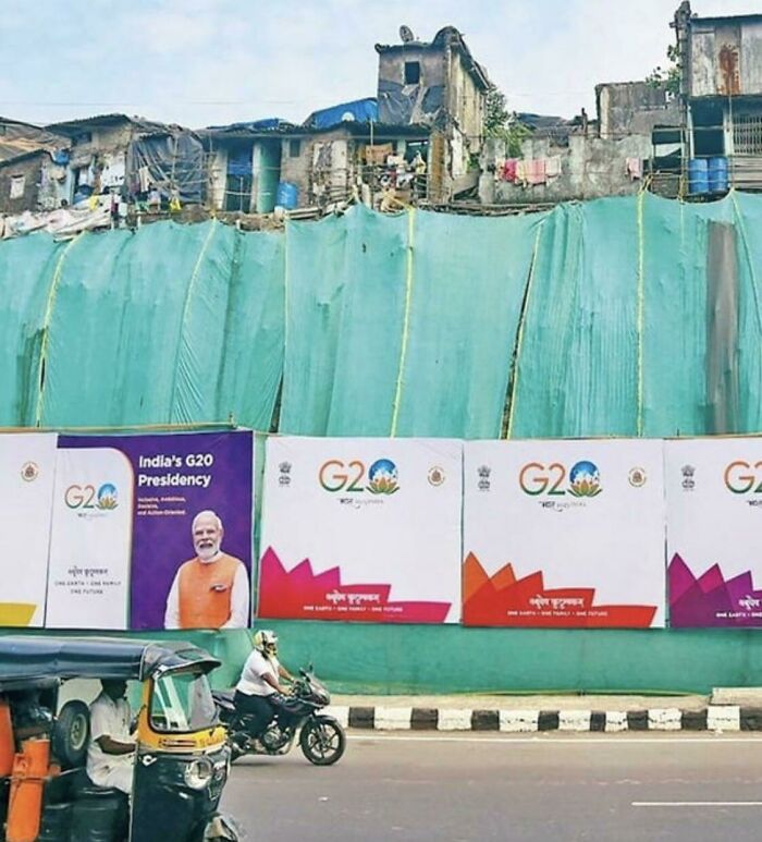 Urban hell scene with dilapidated buildings hidden behind G20 posters, contrasting visuals of neglect and global events.