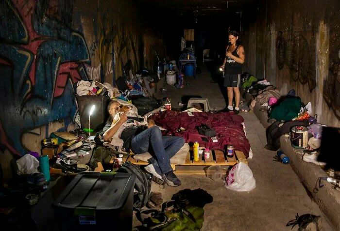 Urban hell example with people living in a graffiti-covered tunnel, surrounded by makeshift bedding and personal items.