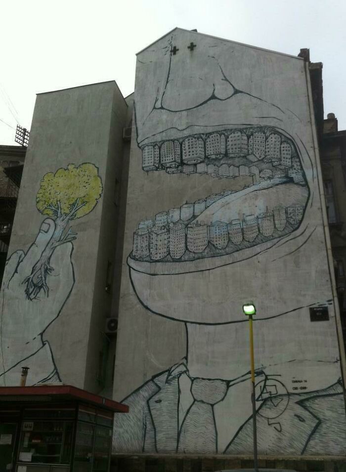 Mural of a large open mouth with buildings as teeth, symbolizing an urban hell theme, on the side of a tall building.