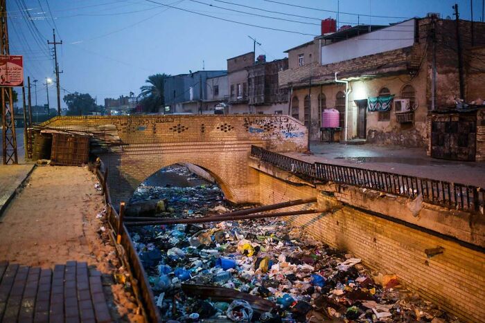 Urban hell example: A polluted canal with trash under a bridge in a run-down city area at dusk.