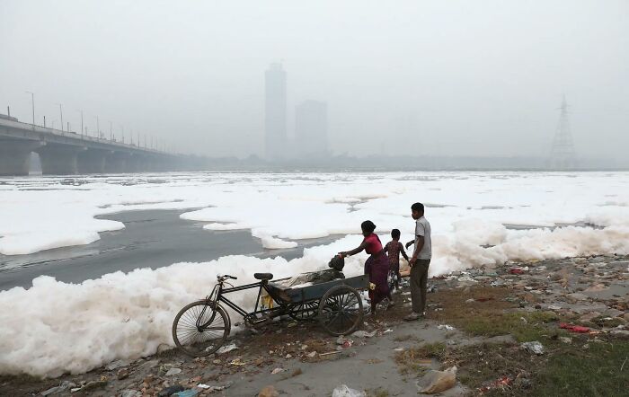 Family by polluted urban river with foamy water, bicycle, and trash on the bank under a hazy gray sky.