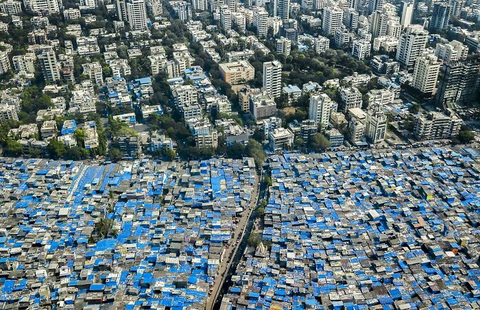 Aerial view of urban hell with dense blue-tarp slums contrasting modern buildings in a cityscape.