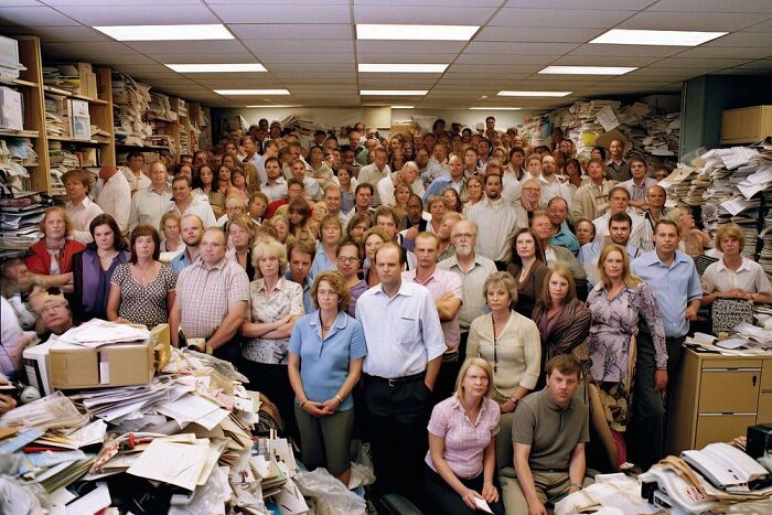 A crowded office with stacks of papers and people standing closely, reflecting symphonic chaos of modernity.