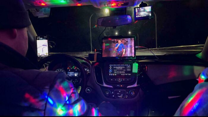 Inside a car at night with decorative lights, devices on the dashboard, and a passenger watching TV; related to Idiots-In-Cars.