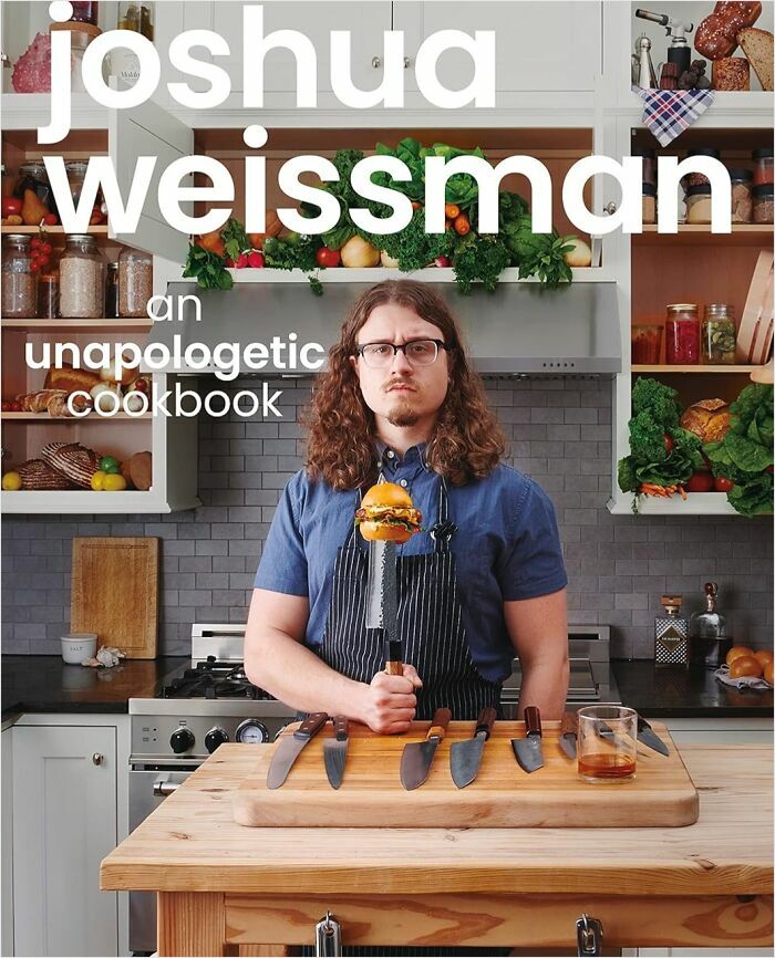 Person in a kitchen holding a burger on a knife, wearing a blue shirt and apron, surrounded by cookbooks and vegetables.