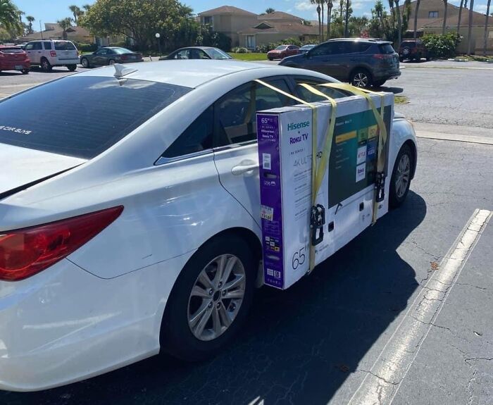 White car with a large TV strapped awkwardly on the side, parked in a lot.